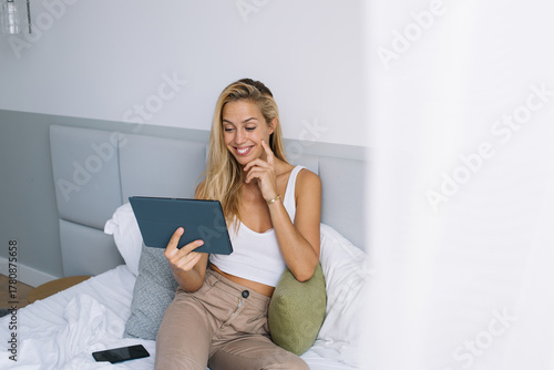 Fotografía Woman smiling and gently touching face while looking at tablet, enjoying digital