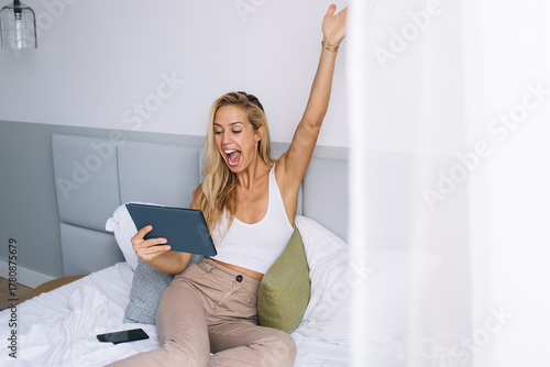 Fotografía Young woman raising hand with joy while looking at tablet, celebrating moment of