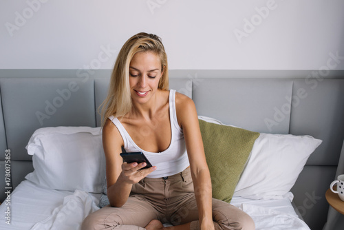 Fotomural Young woman looking at smartphone with a soft smile while sitting in bed, embody