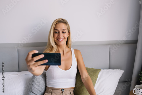 Fotografía Smiling woman holding smartphone at eye level, taking selfie while seated in bed