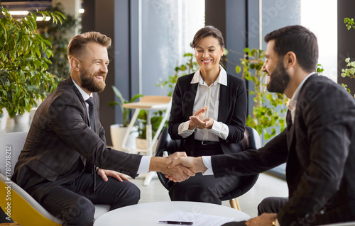 Group of successful company employees making deal sitting in modern office. Happy smiling business people in formal clothes shaking hands on meeting. Teamwork and partnership concept.