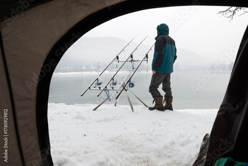 Fishing adventures. Camping and carp fishing with snow in winter on lake. Man near fishing gear covered in snow. Bad weather and cold conditions