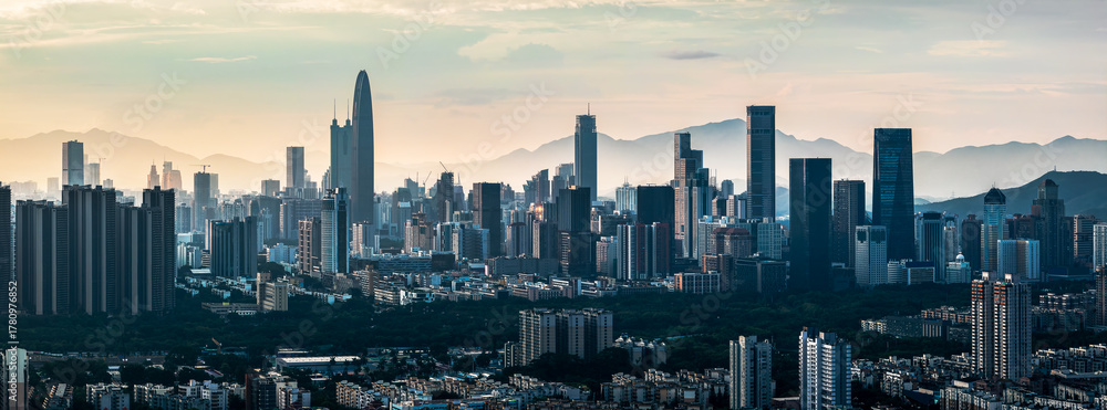 Naklejka premium Telephoto view of the modern city skyline with skyscrapers silhouetted against a hazy mountain range at sunset in Shenzhen.