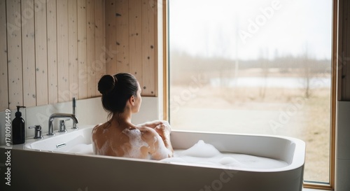 Serene Bath Time in a Minimalist Bathroom. Woman Relaxing in a Bathtub with Bubbles. Tranquil Bathroom Interior with Natural Light.