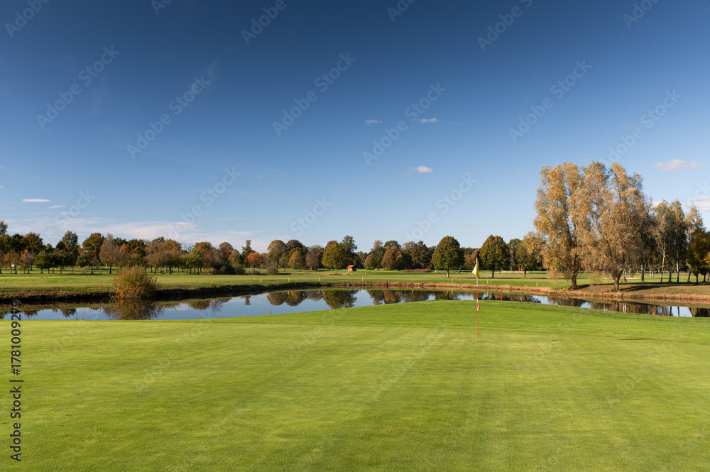 Obraz premium Golf green with flag and pond surrounded by autumn trees