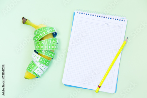 Blank notebook, tape measure and banana on light green background.