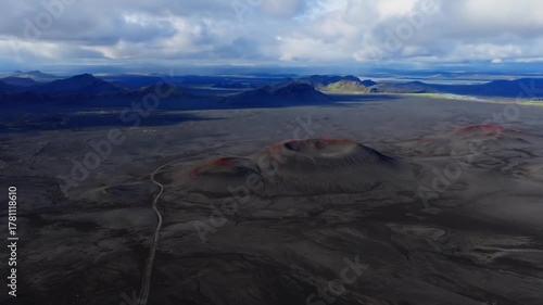 Aerial view shows red rim cinder cones on black lava and ash, a winding dirt road, and distant mountains and lakes in Iceland, under soft daylight with slow pan.