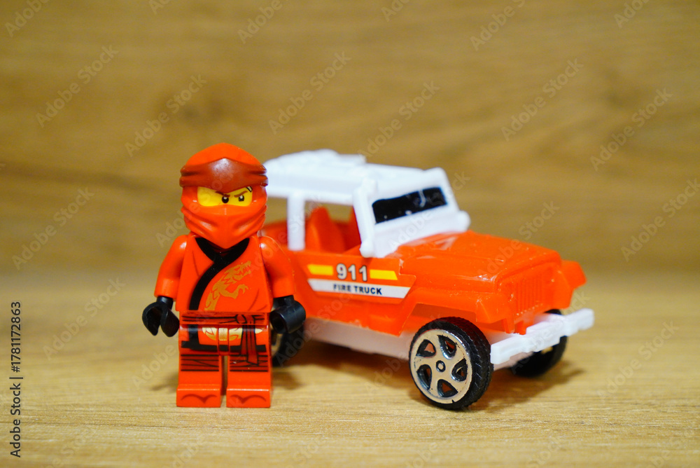 Obraz premium Lego Ninjago minifigure stands next to a toy fire truck on a wooden surface