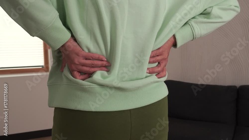 Mature woman touching her lower back in pain while standing at home. Concept of backache, muscle tension, spine discomfort, healthcare, injury and physical therapy recovery.