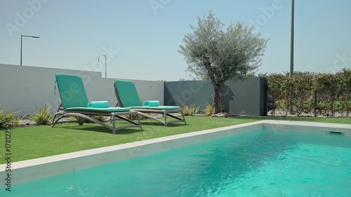 Turquoise water sparkles in a modern swimming pool, bordered by a pristine white deck and artificial turf, with two empty deck chairs inviting guests to unwind and enjoy the sunny day