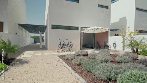 Contemporary white villa with gravel pathway, parked bicycles, patio umbrella, surrounded by lush landscaping creating peaceful residential atmosphere