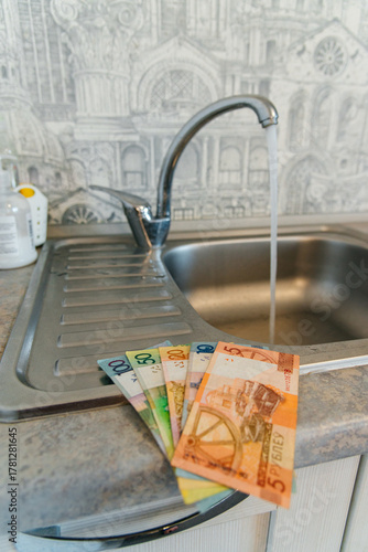 Belarusian rubles at an open water tap in the kitchen, water savings, and increased utility bills