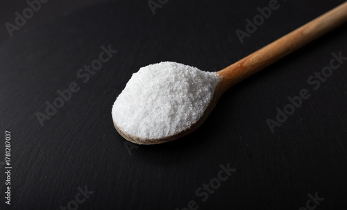 Iodated salt scoop in front of black background with wooden spoon. Textured horizontal photo, closeup. V32.