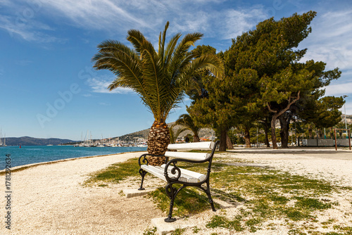 Fototapeta Naklejka Na Ścianę i Meble -  Seaside promenade with palm tree and bench in Trogir Croatia, peaceful coastal view with marina and blue Adriatic Sea