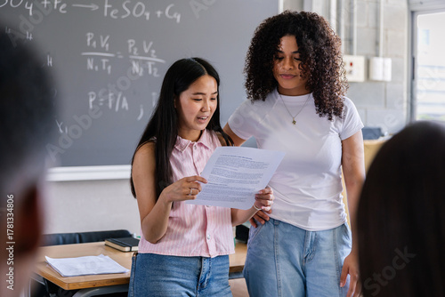 Diverse female students giving class presentation in high school classroom. Two young women speaking confidently in front of classmates during lesson. Education and communication concept.