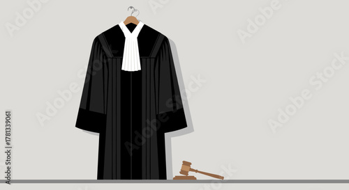 Black judge's robe with white neck band on hanger next to a wooden gavel on a gray surface.