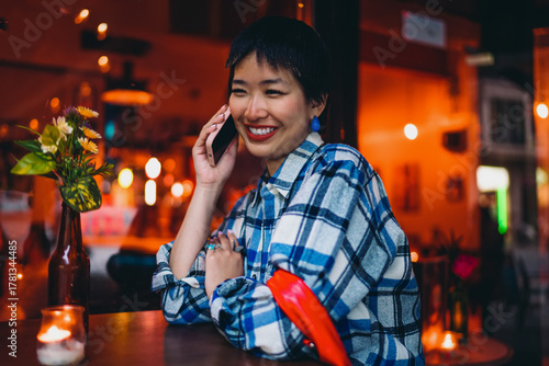 Fotografie Stylish young woman listens during phone call with warm smile, showcasing creative energy, independent spirit and modern tech connection in ambient cafe setting