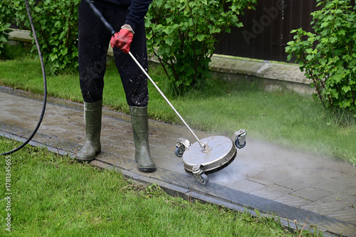 Person using a power washer to clean a stone walkway in a garden setting during daylight hours