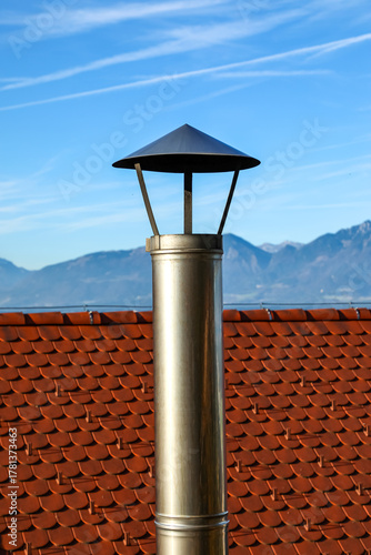 Set against the stunning Alpine mountains, a shiny metal chimney rises from a red tiled roof under a clear sky. The gleaming flue with a round cap symbolizes modern heating and ventilation.