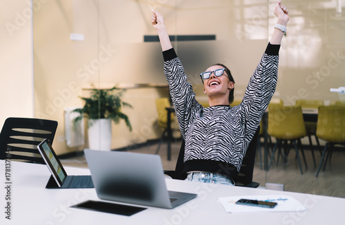 Smiling freelancer raises arms in triumph after completing task, representing confidence, digital independence and pride in solo tech achievements in creative work culture.