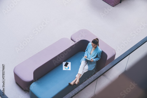Freelancer woman in blue blazer relaxing on pastel sofa with legs crossed, reading digital content on tablet, symbolizing flexible work culture and independent tech lifestyle.