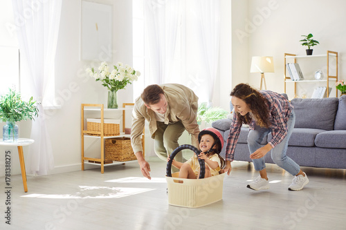 Happy family playing together, parents pushing daughter sitting inside box with steering wheel to pretend driving car. People play in living room at home, child in driver helmet riding laundry basket