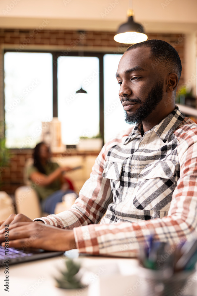 Obraz premium African american male manager typing on laptop, remotely replying to office emails and updating financial reports. Focused black man blogging online using digital device in brick wall apartment.