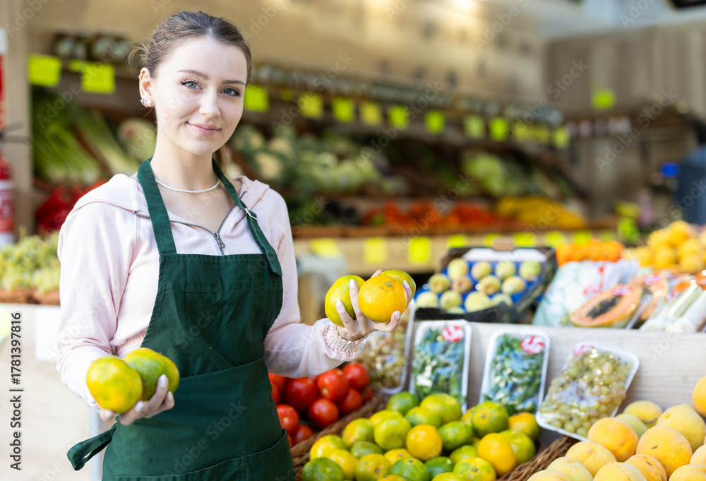 Obraz premium Young woman seller holds ripe tangerines in hands and puts them in a basket on the counter of the vegetable department in the supermarket. Fruit shop worker putting fruit for sale