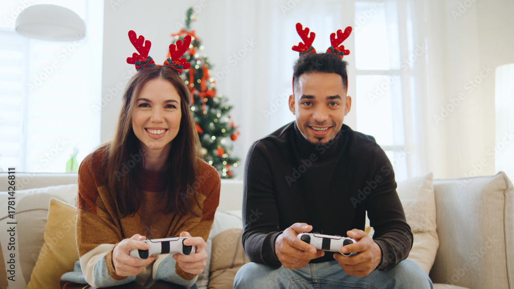 Obraz premium A cheerful couple wears reindeer antlers while playing video games together in their living room, celebrating Christmas and New Year with festive decor around them.