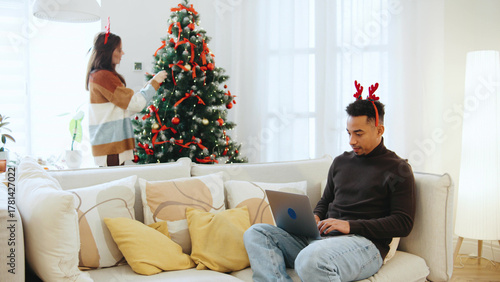 Multiracial couple celebrates Christmas as woman decorates a festive tree while man works on laptop in cozy home setting