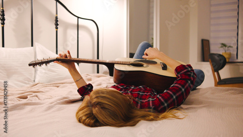A person lies on a bed in a stylish bedroom, casually strumming an acoustic guitar, embodying relaxation and creativity while enjoying some personal time.