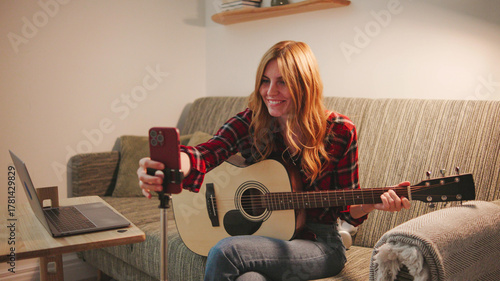 Red-haired young woman in red checkered shirt broadcasts online for blog and plays guitar
