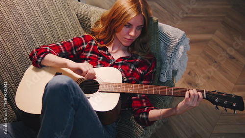 Red-haired Woman playing acoustic guitar in cozy living room with warm lighting and relaxed atmosphere during evening hours