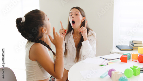 A speech therapist works with a young girl, both animatedly practicing speech and vocal exercises at a bright, modern table filled with colorful learning materials.