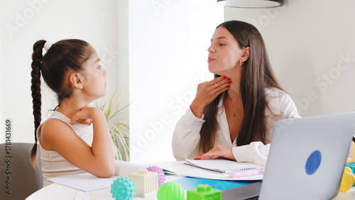 Speech therapist engages with a young girl during a session focused on communication skills, using laptop in a bright office environment