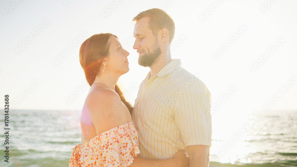 Fototapeta premium Two people stand close together at the beach, gazing into each other's eyes as the sun sets over the water, creating a warm and romantic atmosphere.