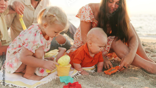 A family spends a joyful time on the beach as three young children play with colorful toys in the sand while the sun sets over the ocean.