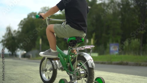 Three year old boy rides a small green bicycle on asphalt in the park surrounded by forest greenery