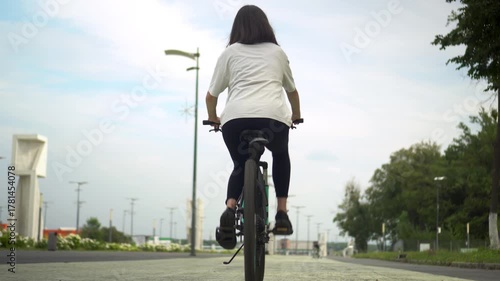 Young woman in white t shirt and black leggings riding bicycle in park rear view 4k