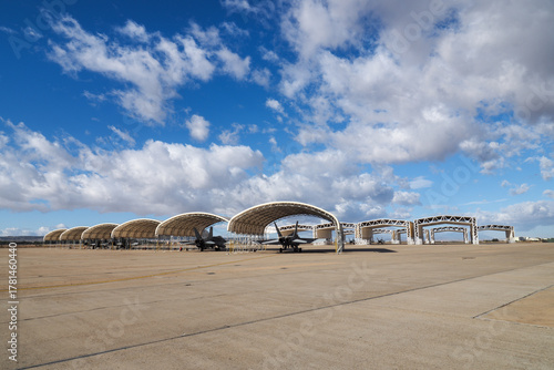 Row of twin engine multirole fighter jets parked under arched sun shelters on a concrete airbase apron under partly cloudy sky