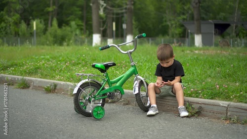 Little boy sits on curb next to kids bicycle with training wheels and looks at his hand