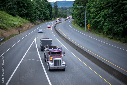 Burgundy big rig tipper semi truck with two tip trailers running on the divided highway road at early morning
