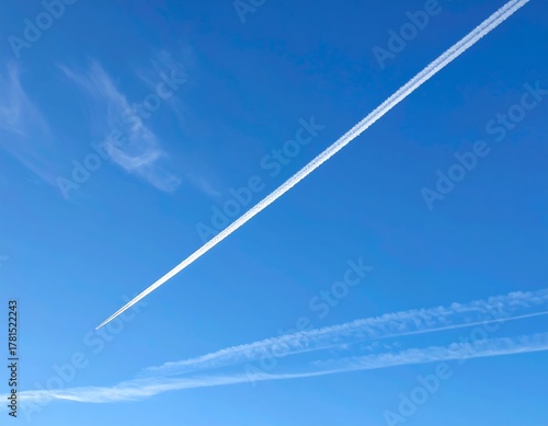 A vibrant blue sky adorned with white contrails created by passing aircraft, forming striking diagonal lines. Delicate cloud formations are also present