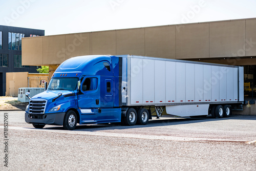 High cab blue big rig semi truck with dry van semi trailer unloading delivered cargo standing in warehouse dock