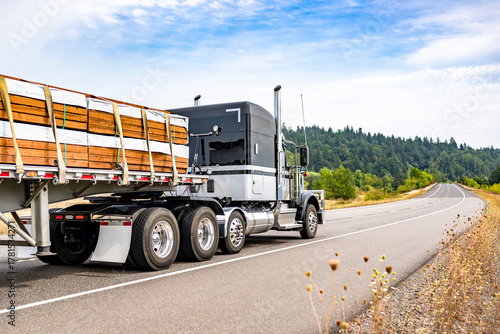 Powerful black and white classic American big rig semi truck tractor transporting fastened lumber wood on flat bed semi trailer driving on the summer winding road