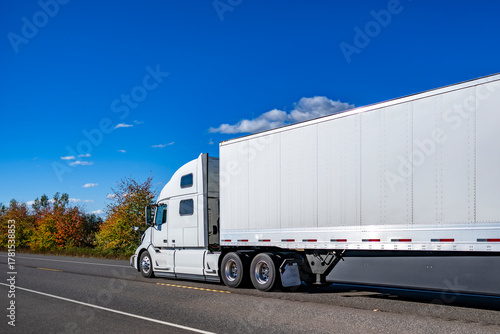 Pro white big rig semi truck tractor with high cab transporting cargo in dry van semi trailer running on the autumn highway road with yellow frees on the side