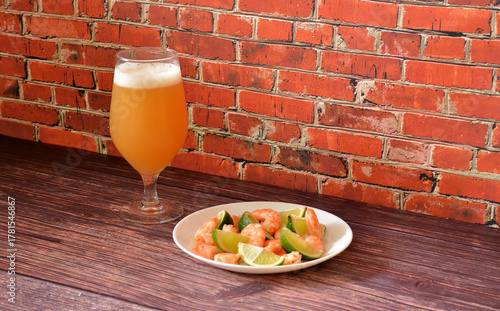 A plate of boiled shrimp with slices of ripe lime and a tall glass of wheat beer on a wooden table.