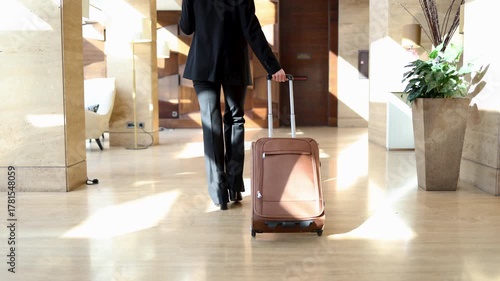 Unrecognizable businesswoman in a formal suit pulling a trolley bag. Rear view of a female traveler walking through a luxury hotel hall