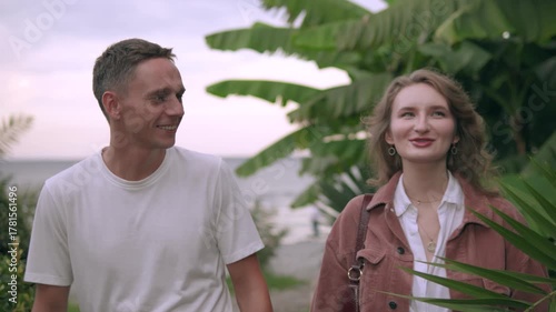 A romantic young couple in their 20s strolls along a tropical seaside at sunset. Palm trees sway gently in the golden light, and the calm blue ocean stretches out before them. 