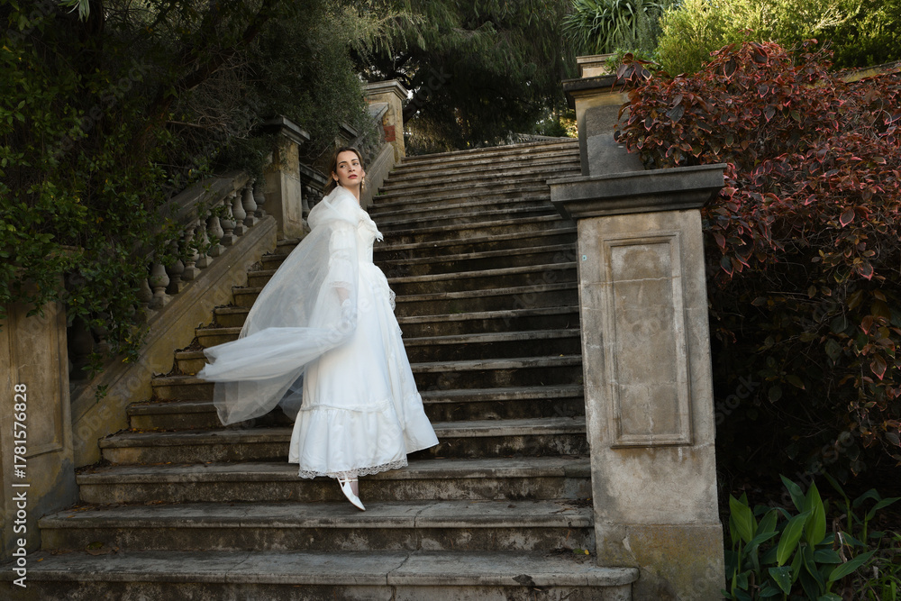 Fototapeta premium portrait of beautiful young blonde model wearing historical white wedding gown and fantasy cloak. wandering ethereal figure walking away on historical staircase of fairytale castle location.